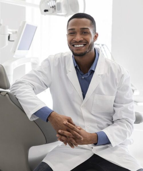 Highly qualified young black dentist posing at clinic over modern cabinet, empty space